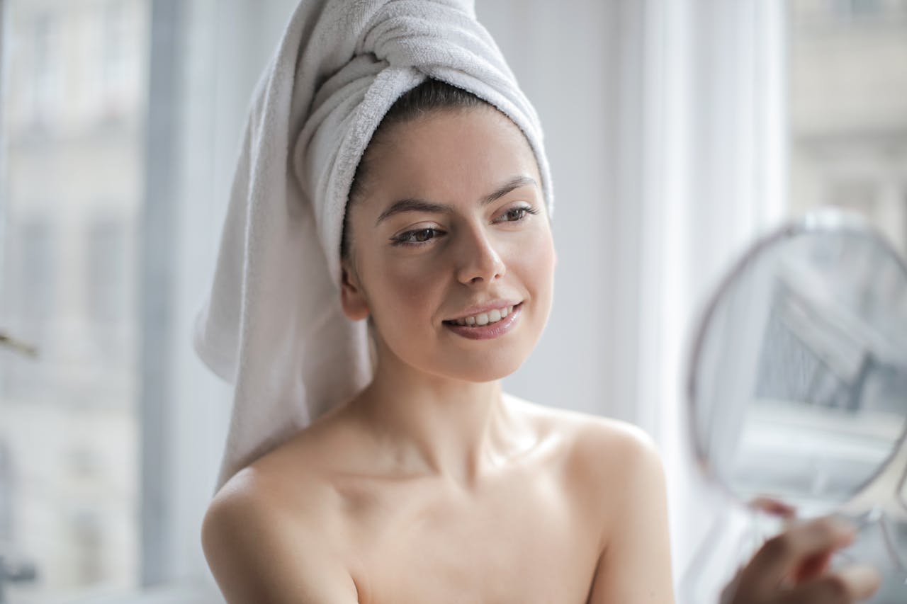 about-us A smiling woman wrapped in a towel looks into a handheld mirror indoors, enjoying her skincare routine.